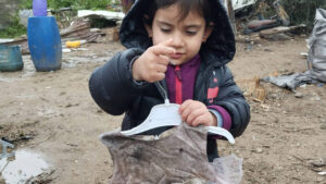 Young girl named Bayan in Gaza, wearing a black hooded jacket and holding up a dirty dress on a hanger while standing in a muddy, makeshift outdoor area.