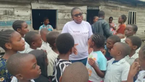 A community leader speaks with Congolese children outside a wooden classroom, highlighting the urgent need for improved education in Eastern Congo.