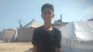 Hani, a 15-year-old orphan in Gaza, standing in front of tents where he lives with his grandmother after losing his parents in a bombing.