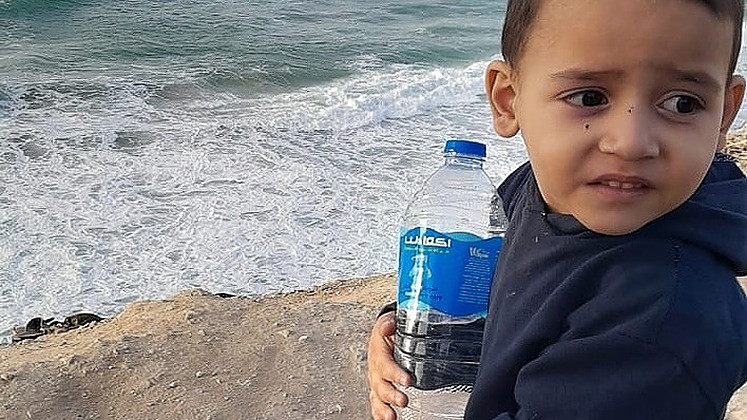 Young boy named Jalal standing by the sea in Gaza, holding a plastic water bottle.