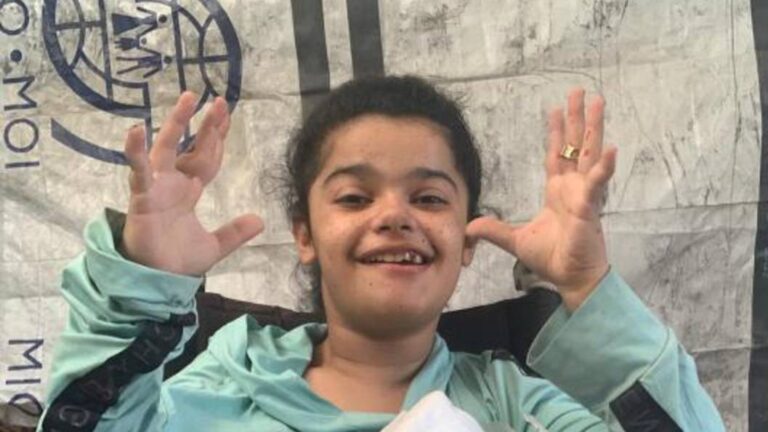 Save Rimas in Gaza – Rimas smiling and raising her hands while sitting in front of a tent.