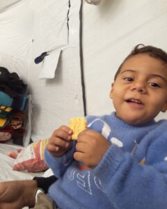 Amer smiling and holding a biscuit inside a tented shelter; used to highlight child nutrition needs for Gaza family emergency aid.