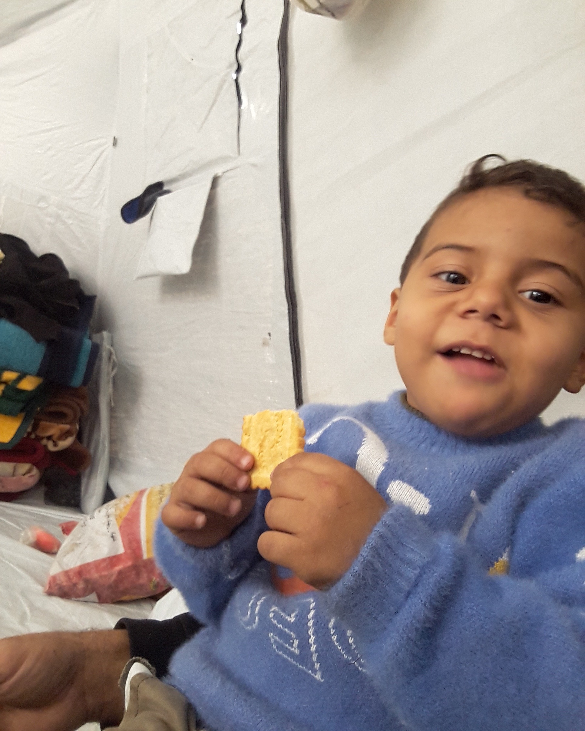 Photo courtesy of Amer’s family. Amer smiling and holding a biscuit inside a tented shelter; used to highlight child nutrition needs for Gaza family emergency aid.
