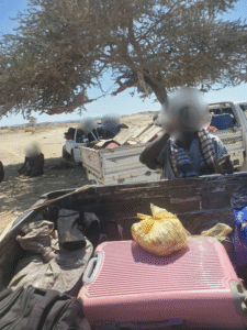 Sudanese student displaced with belongings packed in truck under tree, fleeing war.