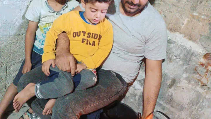 Tariq sitting with his two young sons in harsh shelter conditions; Help Gaza family evacuate.