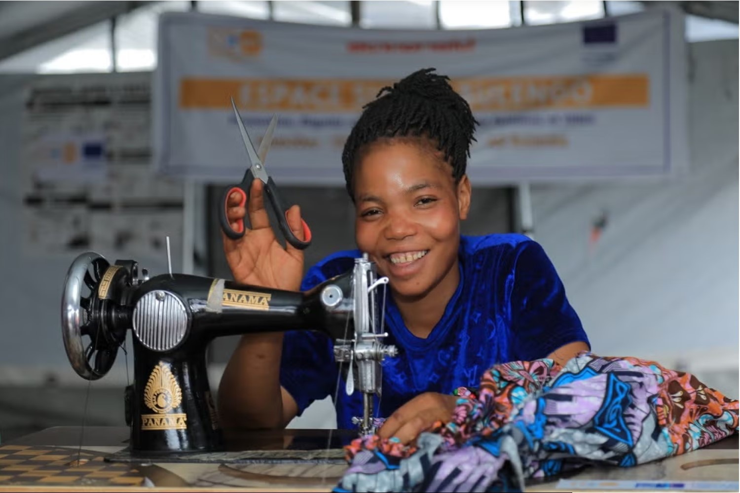 A Congolese woman smiling while holding scissors beside a sewing machine and colourful fabric.