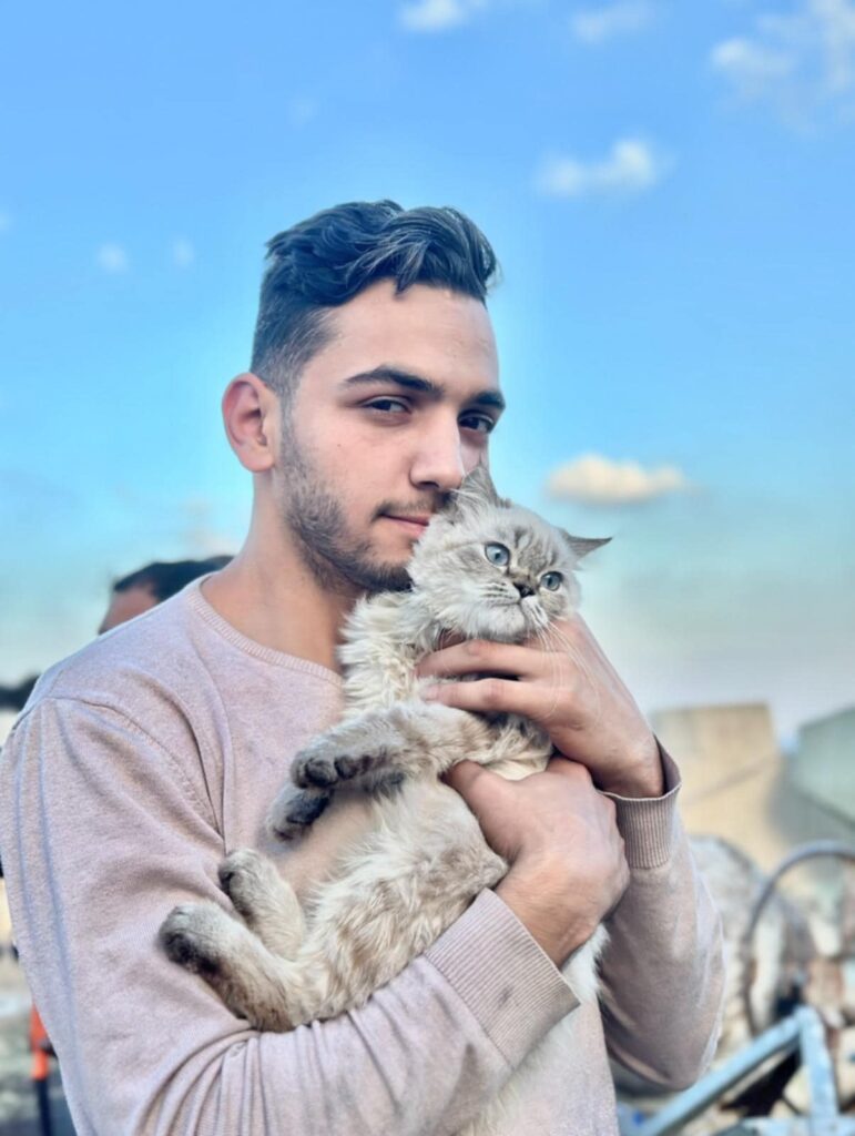 Fares ElMadhoun gently holding his fluffy grey cat against a clear Gaza sky.